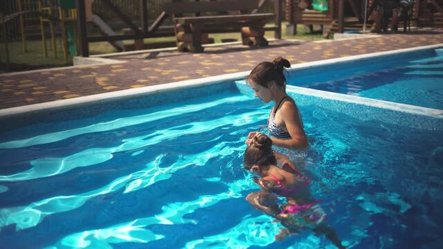 Elder sister in leopard swimsuit pulls little sister out of the pool with blue water