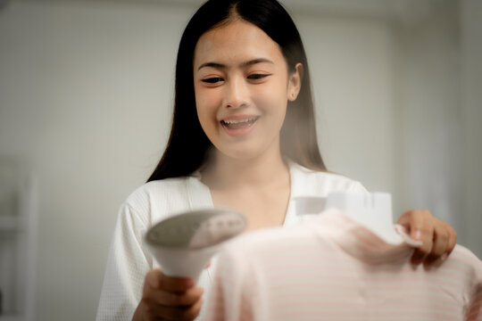 Young Asian woman using handheld garment steamer on baby clothes at home, gentle fabric care, hygiene and sanitation concept, household routine, wrinkle removal and clothing maintenance