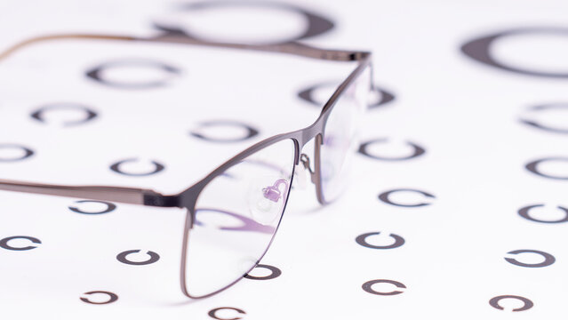Metal eyeglasses rest on an eye chart. Clear lenses gleam softly. Black circles form a vision test pattern. White background emphasizes medical accuracy. Perfect for eye health content
