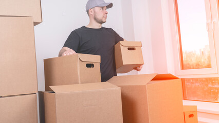 Fototapeta premium Man wears a gray cap and dark t-shirt. He holds a cardboard box. Other boxes stack around him. Natural light streams through a window. His focused expression shows determination