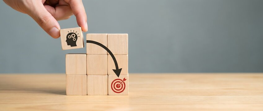 Hand carefully places a wooden block with a cogwheel mind icon on a path leading to a target.