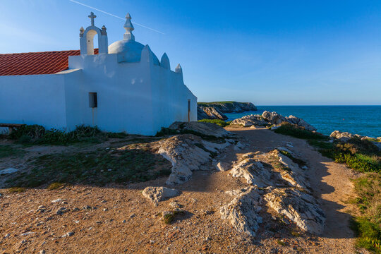 A traditional white coastal chapel with an orange tiled roof and a small bell tower sits on a rocky, sandy terrain