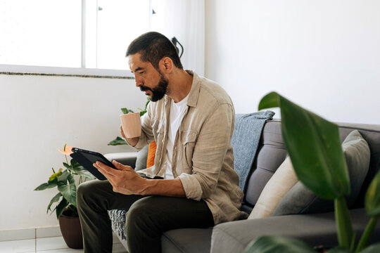 Man sitting on sofa at home using tablet while drinking coffee, relaxing and browsing content in a bright and cozy living room.