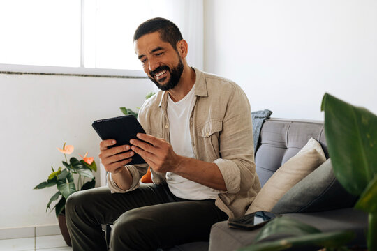 Man sitting on sofa at home using tablet, browsing, reading or communicating in a bright living room environment with natural light.