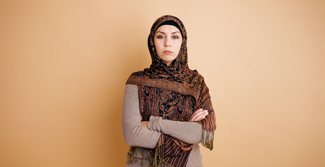 Muslim woman standing with arms crossed, wearing a dark patterned hijab against a peach background,...