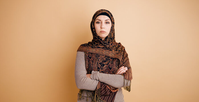 Muslim woman standing with arms crossed, wearing a dark patterned hijab against a peach background, showing seriousness