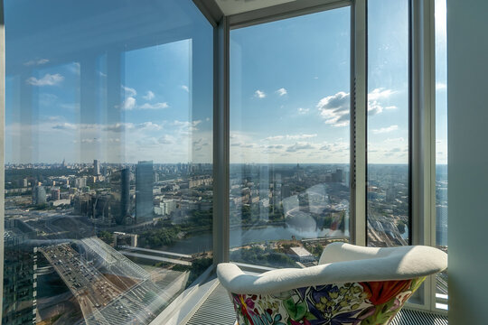 People can see the city landscape from a tall building. The view shows rivers, roads, and distant buildings under a bright sky. A chair adds comfort to the scene