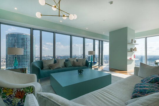A living room featuring a large window offering a view of the city skyline. Bright light enters the room. Furniture includes a blue coffee table and sofas