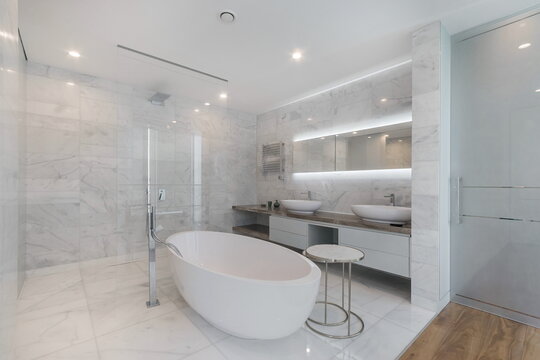 A sleek bathroom shows a freestanding tub, two sinks, and large mirrors. Modern lighting shines on white marble walls and floor, creating a clean look