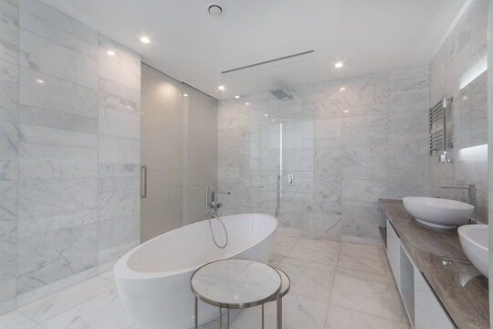 A bathroom features a freestanding bathtub and two sinks situated on a wooden countertop. The wall is covered in white marble tiles, and bright lights enhance the space