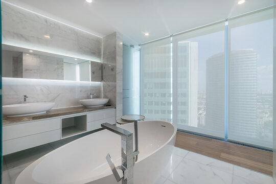 A bright bathroom features large windows that show a city view. The design includes a tub and double sinks. Light reflects off the marble surfaces, offering a stylish space for relaxation