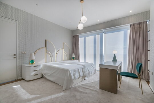 A modern bedroom is shown with a large bed and simple decor. The room features large windows that let in sunlight and a stylish desk and chair