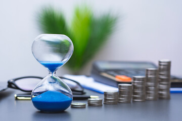 Hourglass with blue sand stands beside stacked coins on an office desk. Concept time for money,and financial decision making. Business planning, investment strategy, productivity and savings. © vadish