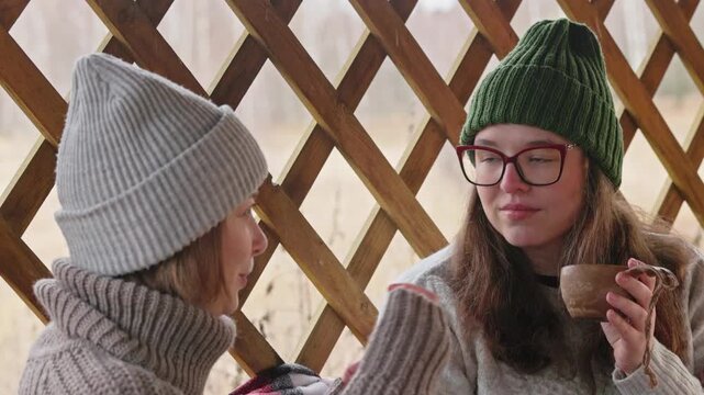 Two friends enjoy hot drinks while chatting and smiling at a cozy cafe on a chilly day, wrapped in sweaters and hats.