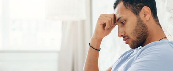Fototapeta premium A man is sitting alone in a room, resting his forehead on his hand. He appears deep in thought, with his gaze directed downwards. The setting is casual and simple, focused on his expression.