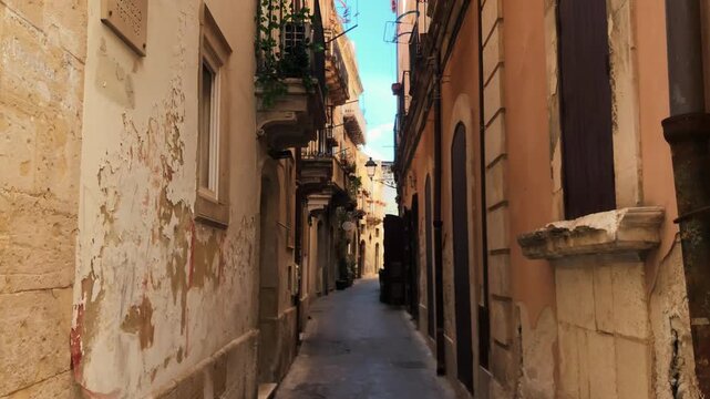 A beautiful view of an empty narrow street lined with old historic buildings featuring charming balconies on a sunny day in the old town of Syracuse, Sicily, Italy.
