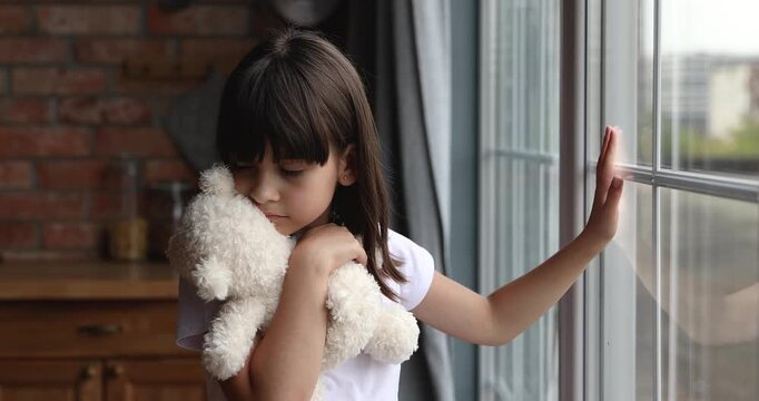 Sad child stands alone near window holding bear toy, looking outside thoughtfully, feeling lonely and upset, childhood reflection, challenges, isolation, need for comfort and emotional support at home