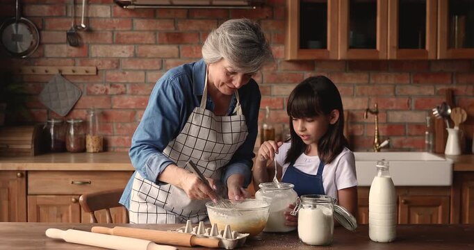 Grandmother teaching child granddaughter to cook, preparing homemade pastry in kitchen, mixing dough, adding flour, enjoy communication and time together. Intergeneration family bonding, home cooking