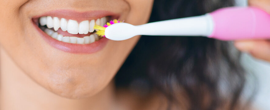 A woman is using an electric toothbrush to clean her teeth while standing in front of a mirror. The setting is a bright bathroom, and she is focused on her oral hygiene.