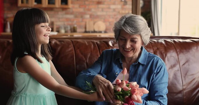Granddaughter congratulate grandmother with birthday, giving her bouquet of tulips flowers, gift box, sharing hugs and kisses, expressing love. Celebration of family holidays, intergenerational bond