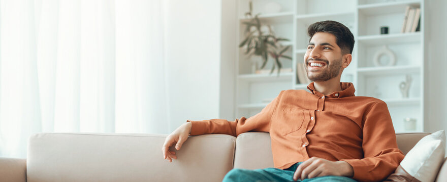 A young man sits comfortably on a couch in a bright room. He has a smile on his face and seems to enjoy his leisure time. Sunlight is streaming through the window, creating a warm atmosphere.