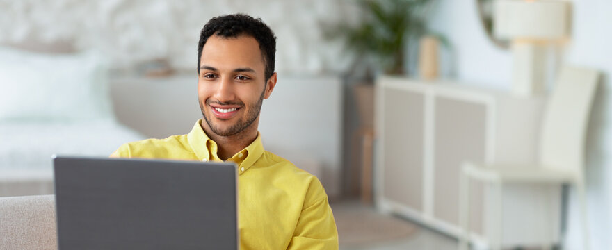 A man sits on a sofa at home, using a laptop. He smiles as he engages with the screen. A bright and modern room is visible in the background with soft colors and furniture.