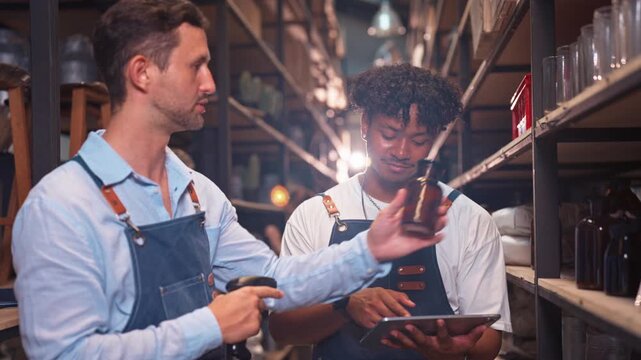 White male and a Black male colleague both wearing aprons study inventory data on a tablet together while standing in a storage warehouse aisle.