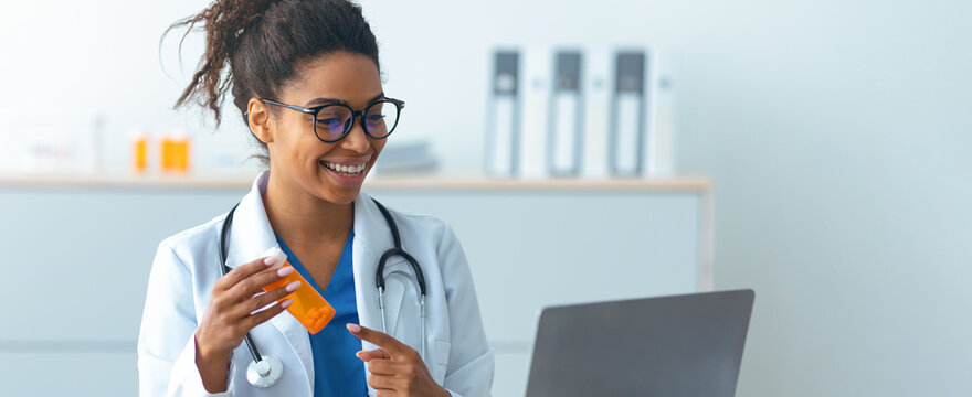 A doctor smiles while holding a prescription bottle and pointing at a laptop screen in a clinic. The doctor is engaged and appears to explain medication details to a patient.