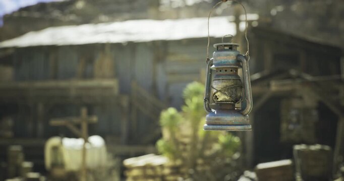 A vintage lantern is suspended in the foreground near an aged building in a remote area. The setting suggests a bygone era, surrounded by wooden structures and natural elements.