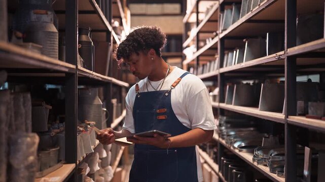 Young Black man in a denim apron carefully checks inventory data on a tablet while standing alone in a long aisle of shelved goods in a warehouse.