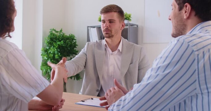 Signing contract for purchase or sale of property by couple during consultation with real estate agent at office. People shake hands to celebrate success of deal, broker and buyers sitting at desk