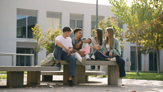 Group of young college students sitting on a bench outside university building, communicating and studying together during break