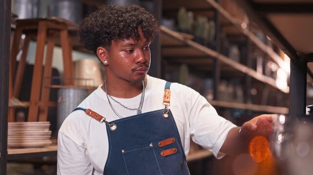 Young Black man in a denim apron holds an amber glass bottle up to examine its contents closely against the warm lighting in a rustic warehouse.