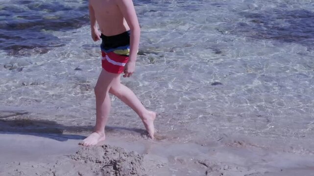 happy child on beach, healthy boy 9-10 years old playing with sand on shore near water mediterranean sea, rotates around himself, creative sand play, foot massage, wonderful vacation, improve health