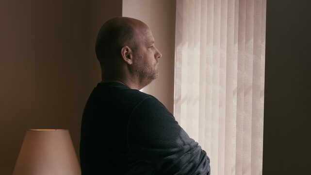 Chubby white man with a somber tone and a shaved head looking out a window with vertical blinds 