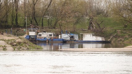Niebiesko-biała barka cumuje przy brzegu rzeki w otoczeniu gęstych drzew. Spokojna woda odbija zarysy maszyn i roślinności. To klasyczny widok śródlądowej żeglugi na tle dzikiej, polskiej przyrody © tomo81