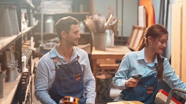 Caucasian man and a smiling Asian woman in mustard yellow aprons review items on a store shelf together inside a bright, cozy homeware retail shop.
