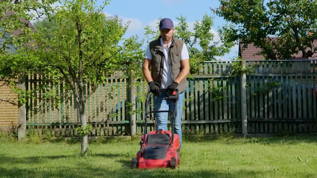 Young man in cap wearing gloves and mowing green grass in the backyard. Gardener pushing lawnmower. Garden work concept. Real time