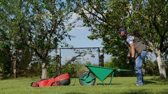 Young man emptying lawnmower grass catcher while pouring grass into a wheelbarrow. Gardening and landscaping concept. Real time