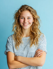Obraz premium Portrait of a smiling teenage girl with long wavy blonde hair and blue eyes. Happy young woman with crossed arms wearing a striped t-shirt, isolated on a bright blue background