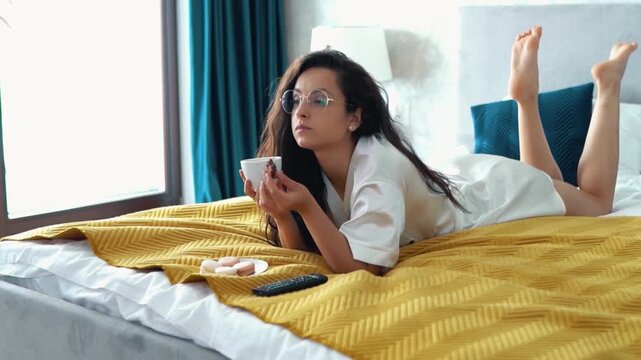 Hot Caucasian girl in glasses and lying on her bed, watching tv with interest, drinking coffee from white cup and eating biscuit. Beautiful woman eating chocolate, and moving her legs in her bedroom.
