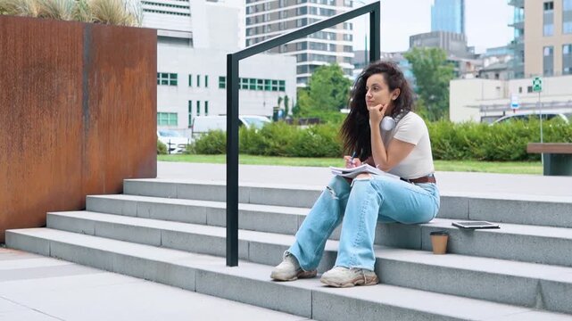 Smiling young lady making notes in notebook while sitting outdoors on steps. Digital tablet and coffee cup on stairs. Education in university college concept. Real time