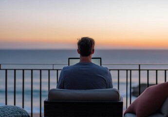 Obraz premium Man focusing on his laptop, working remotely from a balcony with an expansive ocean view during a calm sunset