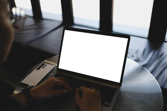An over the shoulder shot of a person typing on a laptop with a blank white mockup screen placed on a round metallic table.