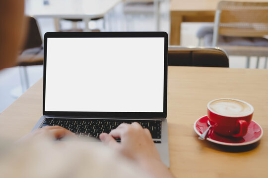 An over the shoulder shot of a person typing on a laptop with a blank white mockup screen placed on a wooden table next to a red cup of coffee.