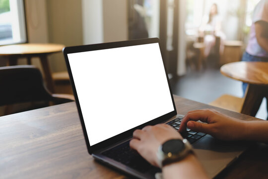 A close-up view of a person typing on a laptop with a blank white mockup screen while sitting at a wooden table in a cafe.