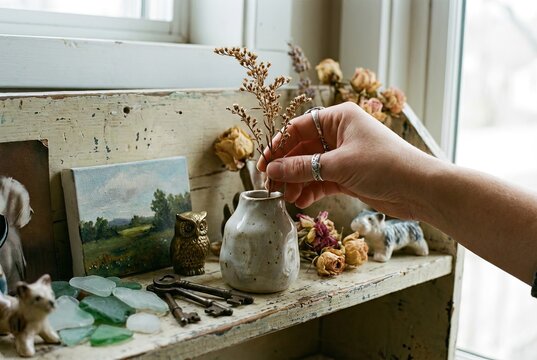 Aesthetic Close-Up of Hand Arranging Decorative Sprig in Ceramic Vase on Curated Wooden Shelf with Personal Treasures