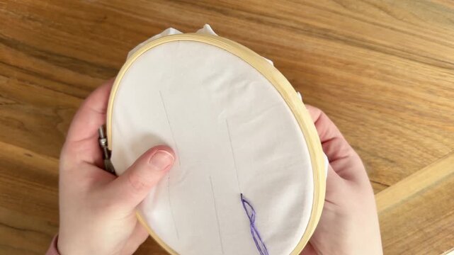 Female Caucasian hands demonstrating a back stitch in an embroidery hoop using purple thread on white fabric. Close-up 4K needlework tutorial showing stitching technique on wooden table.