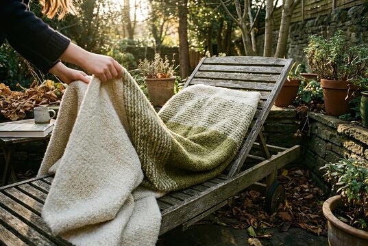 Hands gently draping a soft blanket over a cozy lounge chair in a peaceful garden nook bathed in warm afternoon sunlight, preparing for ultimate relaxation