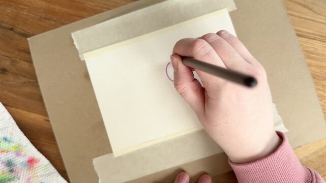 Female Caucasian hand painting a purple circle on dry watercolor paper, demonstrating wet-on-dry technique with clean edges. Close up 4K painting process on taped paper.
